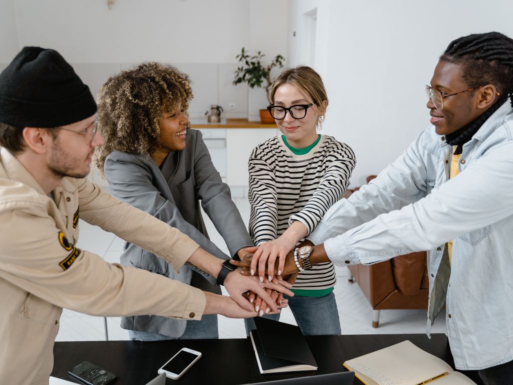 Four people placing their hands together in a gesture of teamwork or unity over a table with devices and notebooks.